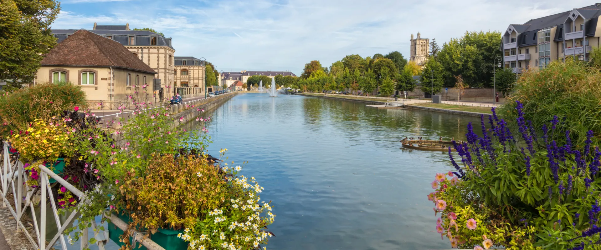 Canal fleuri à Troyes avec bâtiments historiques et végétation colorée bordant l'eau calme.