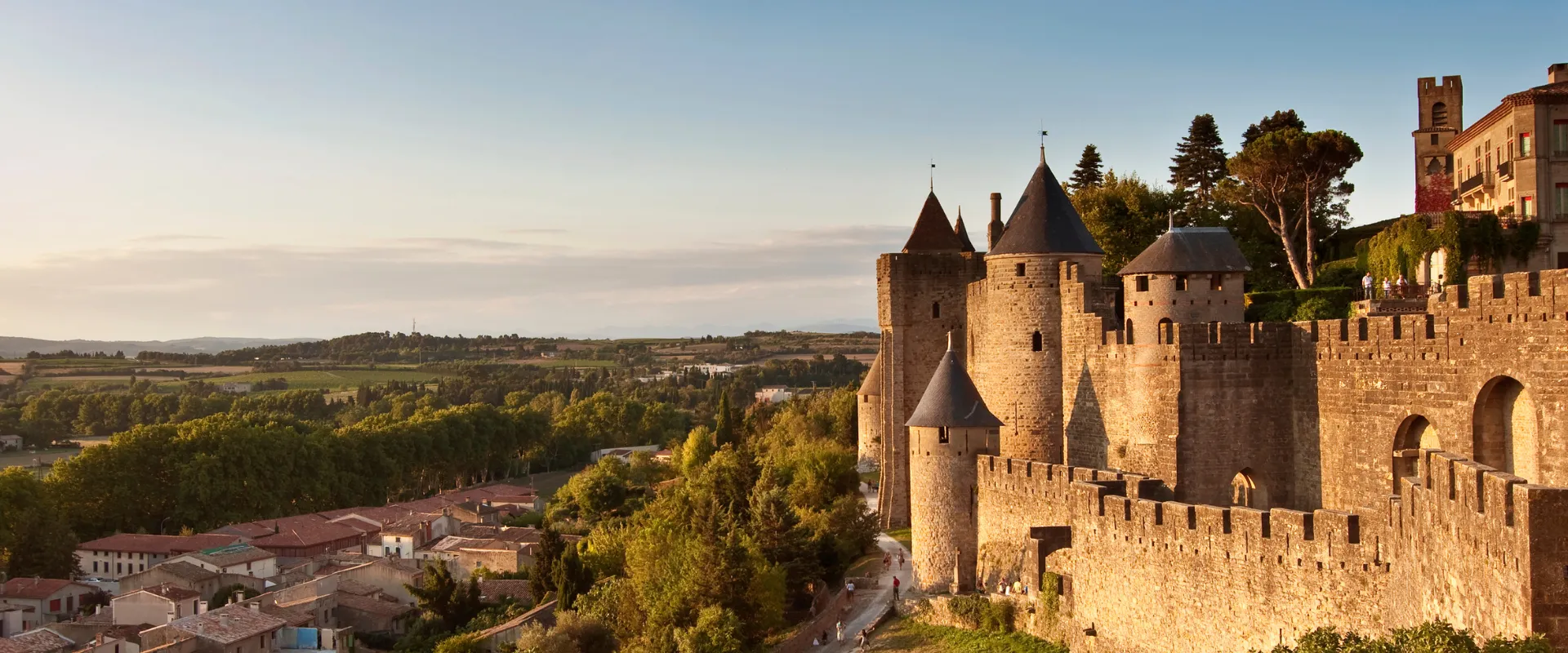 Forteresse médiévale d'Aude avec tours rondes et crénelages dominant vallée verdoyante.