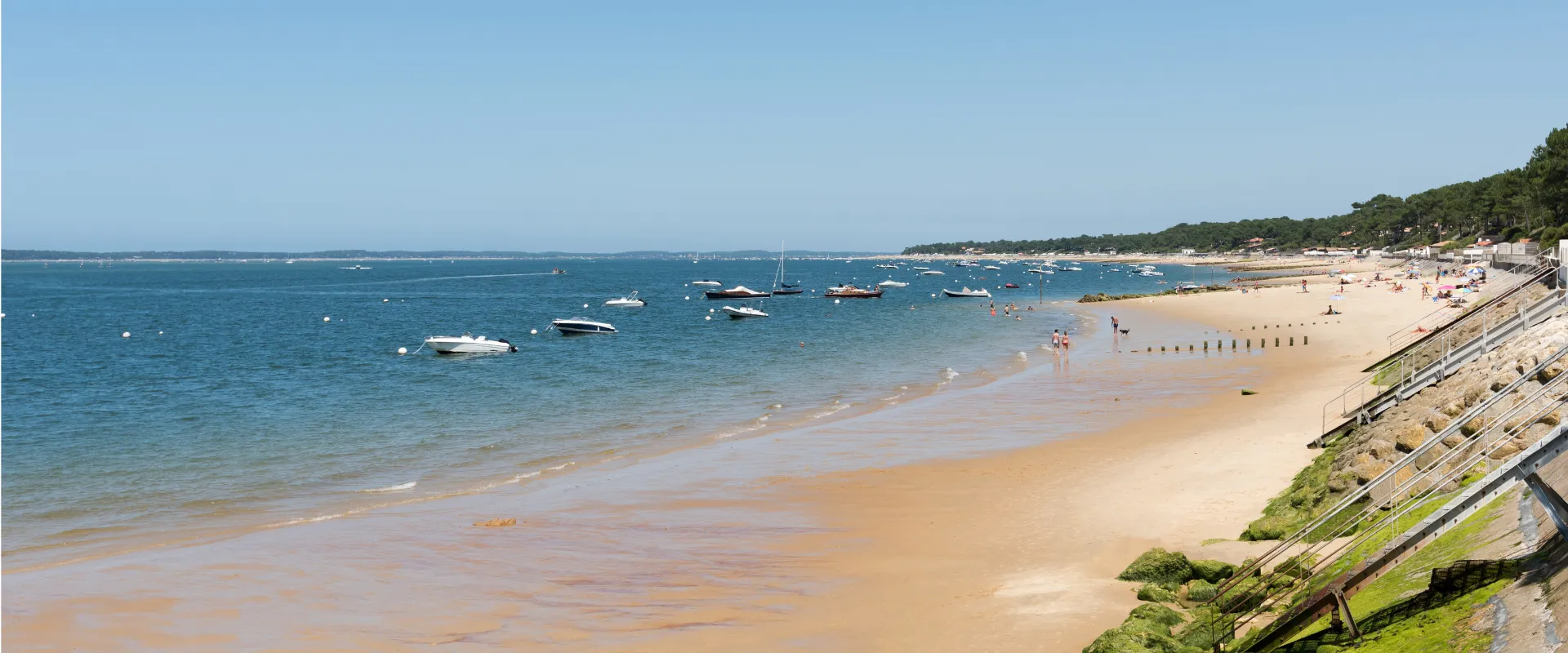 Plage de sable fin avec bateaux ancrés et falaises couvertes de végétation verdoyante.