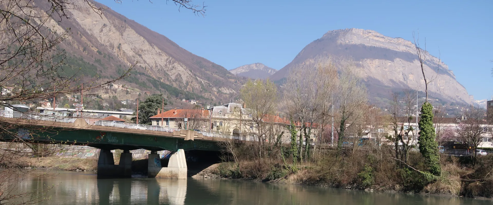 Pont sur rivière avec montagnes boisées et villages alpins en arrière-plan en Isère.