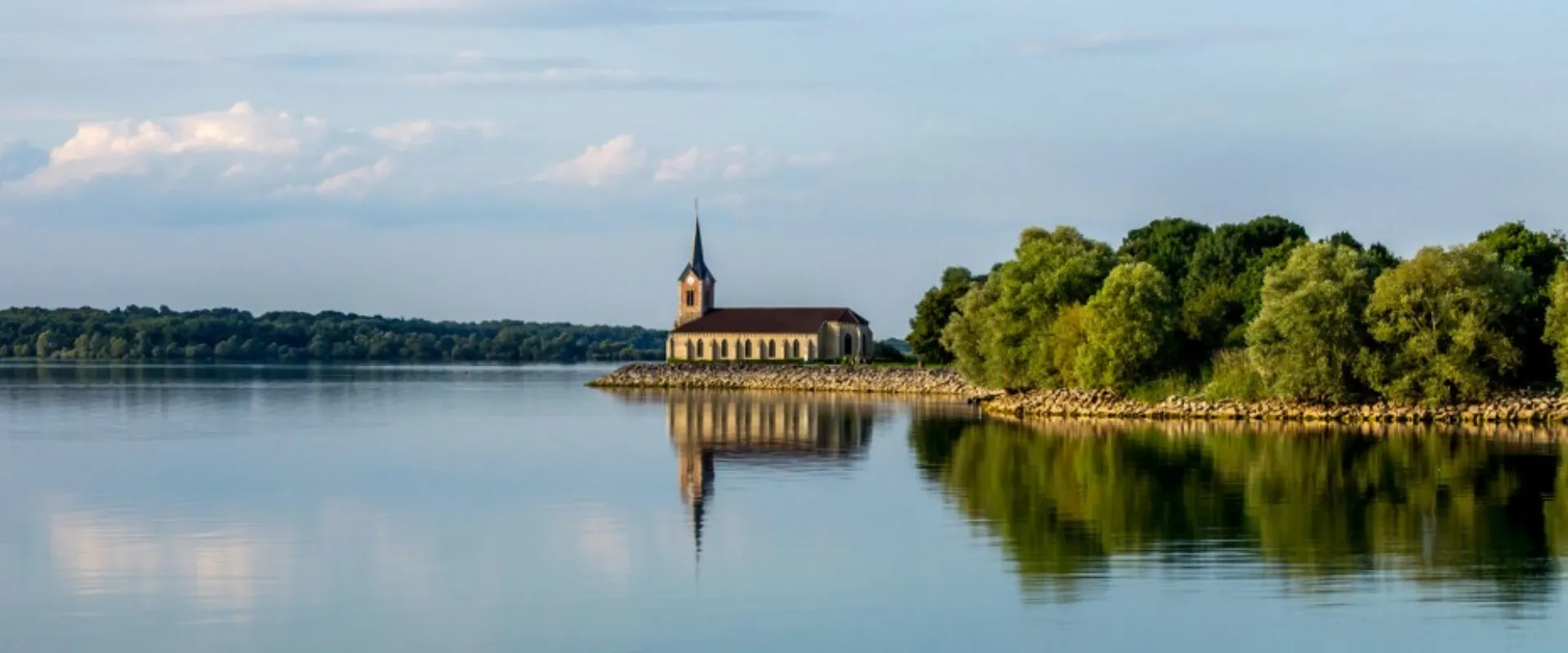 Église en pierre avec clocher noir au bord du lac calme, entourée de végétation verte.