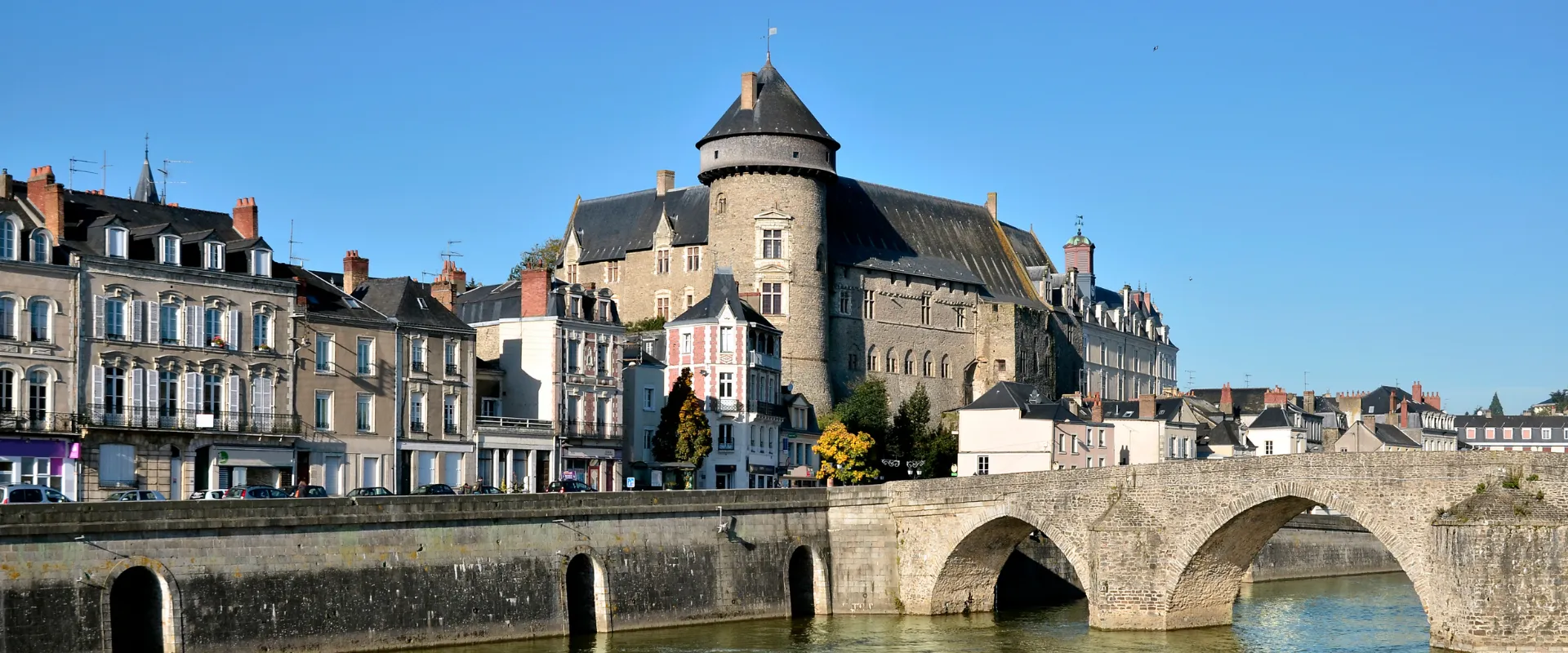 Château médiéval avec tour ronde dominant ville historique et pont de pierre sur rivière.