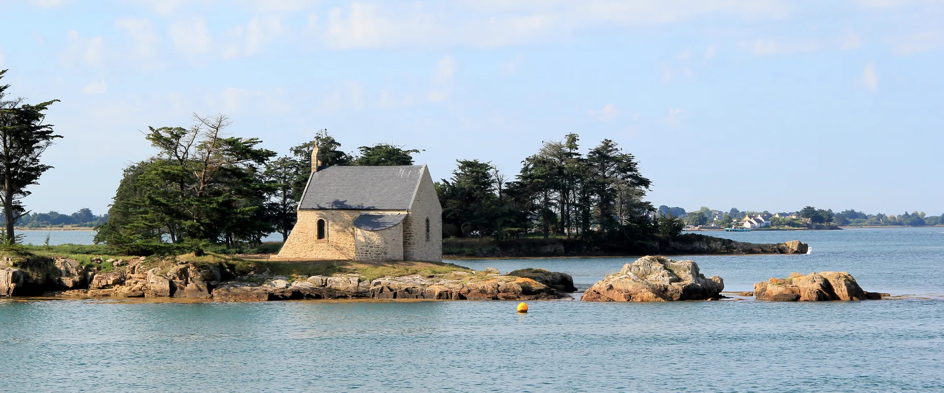 Chapelle de pierre isolée sur îlot rocheux en Morbihan avec arbres et mer turquoise.