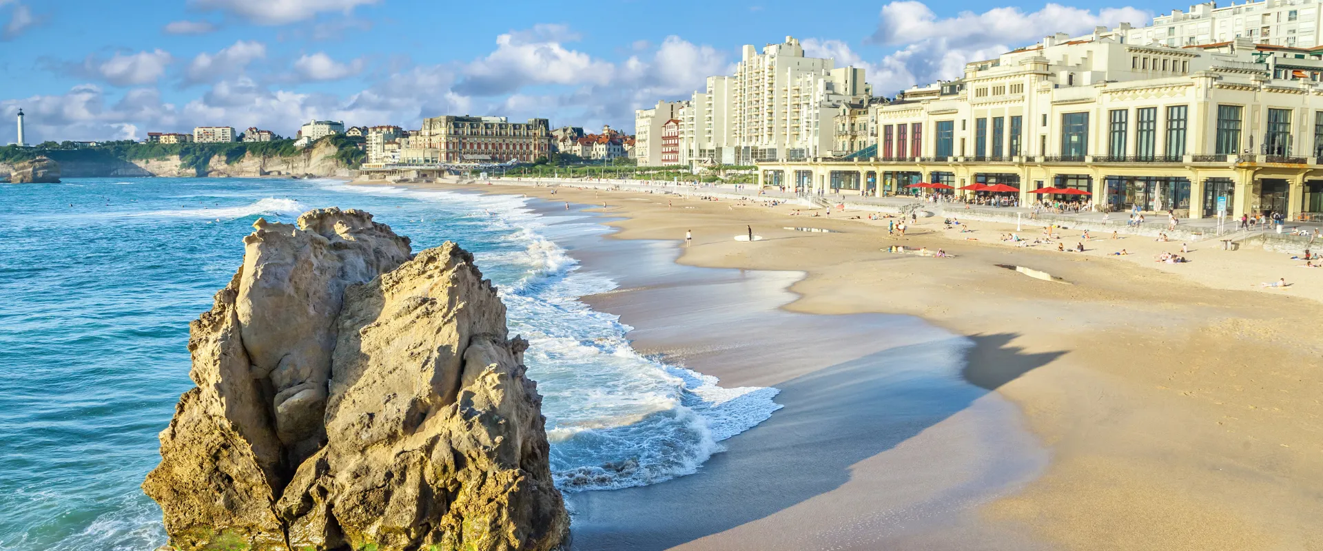 Plage de Biarritz avec rocher, vagues, sable doré et bâtiments historiques côtiers.