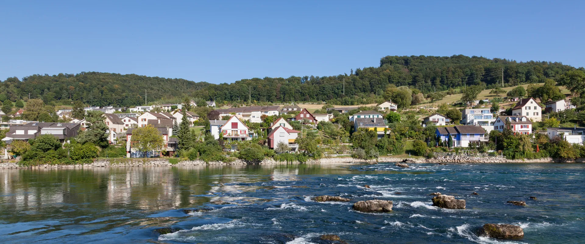 Village alsacien au bord du Rhin avec maisons colorées et forêts verdoyantes.