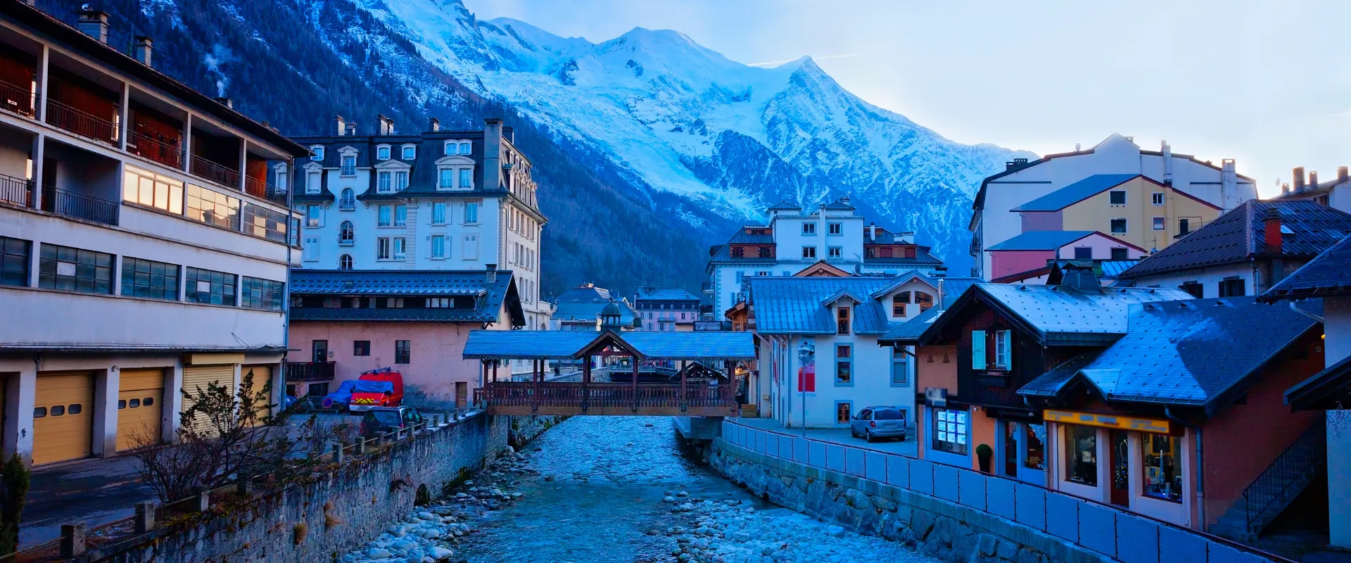 Village alpin enneigé avec rivière, bâtiments colorés et montagnes bleutées en arrière-plan.