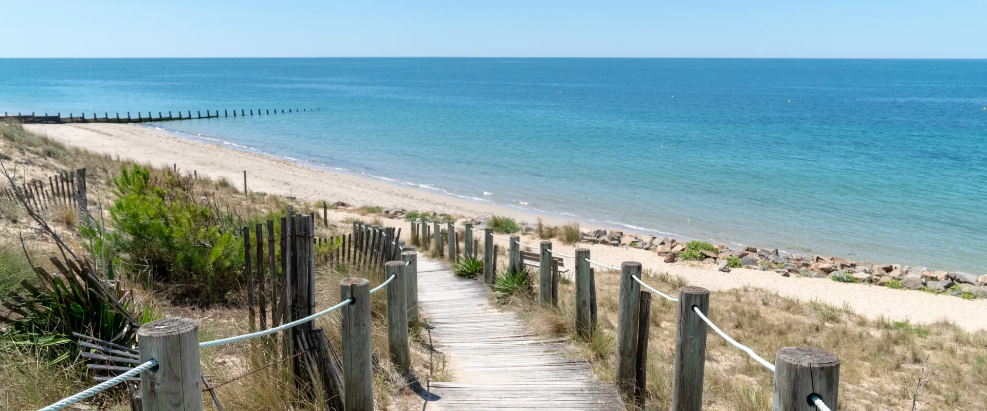 Boardwalk en bois menant à plage de sable fin avec dunes herbeuses et mer turquoise Vendée.